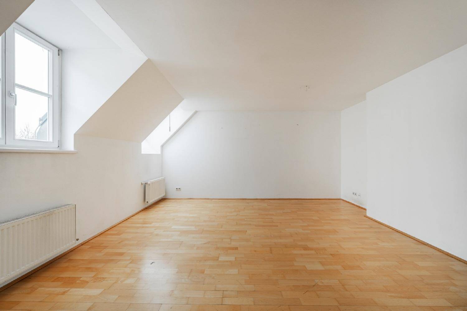 Geräumiges Zimmer mit Holzparkett, weißen Wänden und einem großen Fenster unter der Dachschräge.