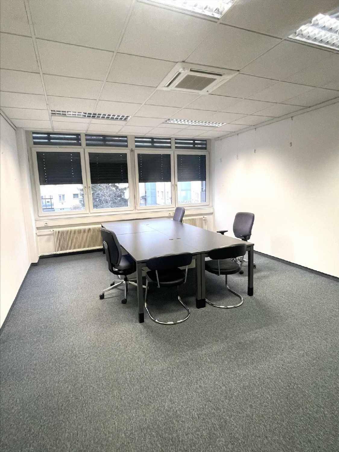 Büro mit dunklem Tisch und Stühlen, grauem Teppichboden und Fenstern mit Jalousien.