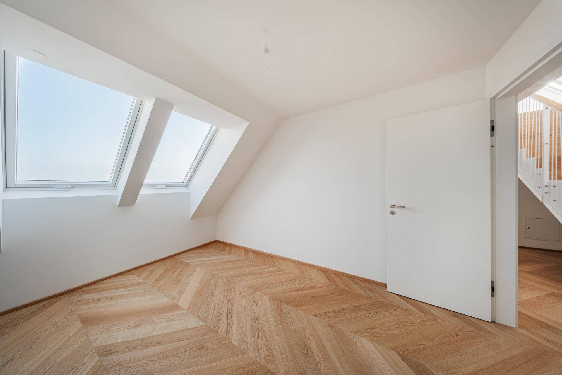 Helles Zimmer im Dachgeschoss mit schräger Decke, Parkettboden und zwei Dachfenstern.
