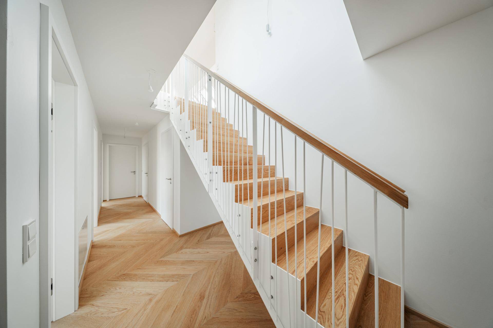 Moderner Treppenaufgang mit Holzstufen und weißem Geländer, der zu einem hellen Flur führt.