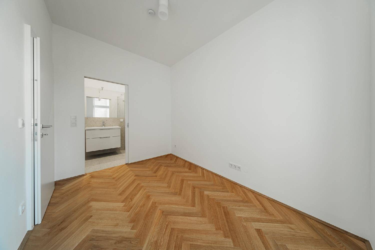 Helles Zimmer mit Fischgrätparkett und Blick auf ein modernes Badezimmer mit Waschtisch.
