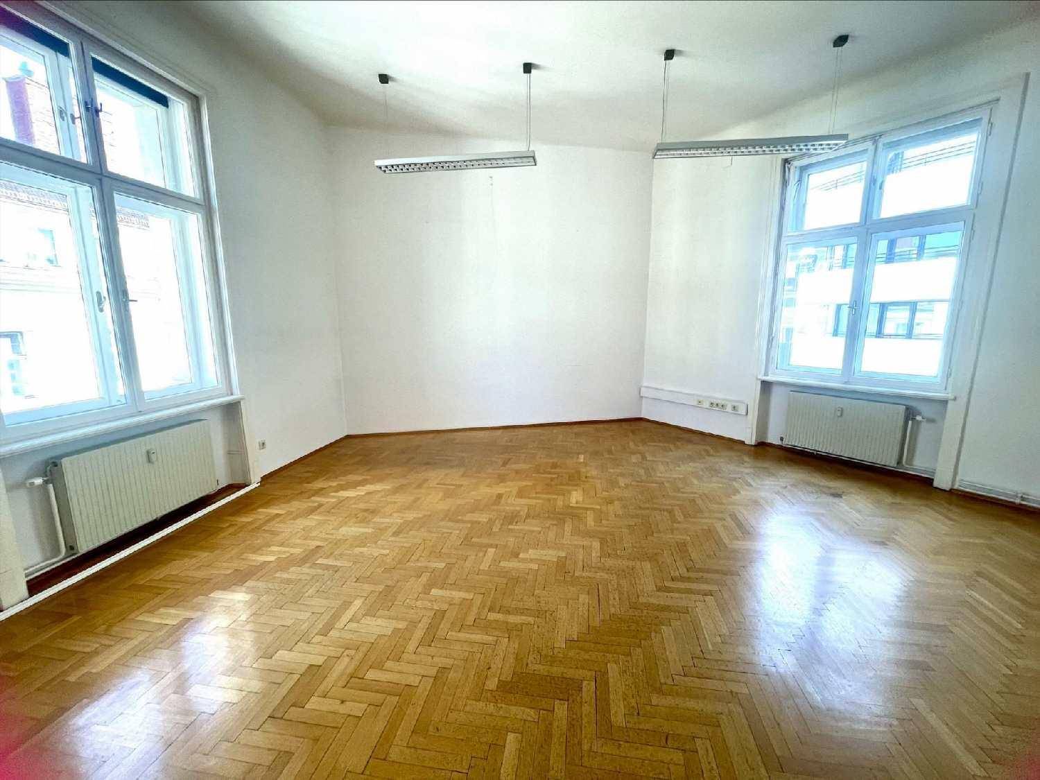 Helles, leeres Büro mit Fischgrätparkett, zwei großen Fenstern und einfacher Deckenbeleuchtung.