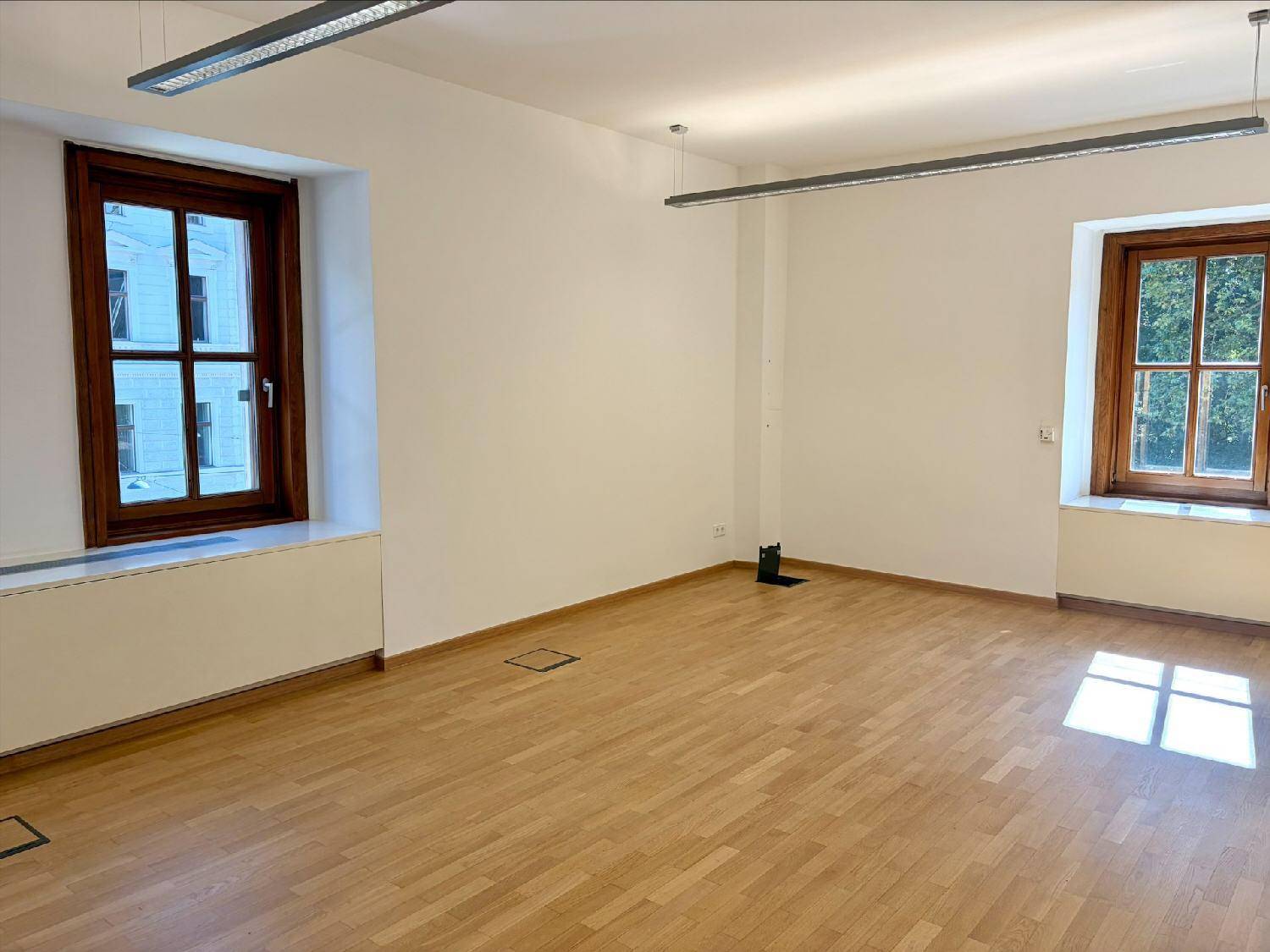 Großer, heller Raum mit Holzboden und weißen Wänden, ausgestattet mit modernen Beleuchtungselementen und Fenstern.