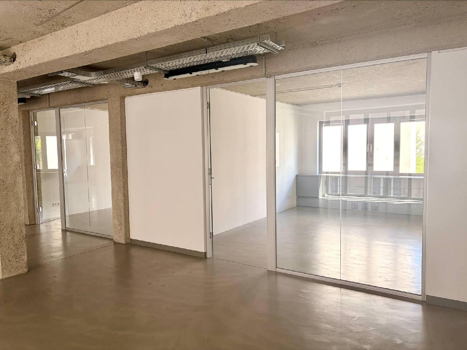 Offener Bürobereich mit Betonboden, Glaswänden und viel Tageslicht durch große Fenster.