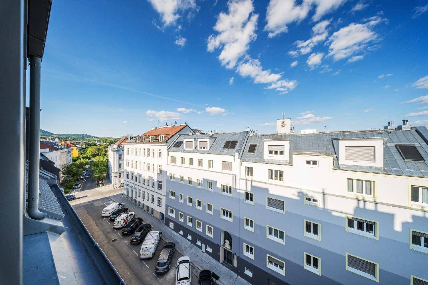 Blick von einem oberen Stockwerk auf eine städtische Straße mit parkenden Autos und umliegenden Gebäuden unter blauem Himmel.