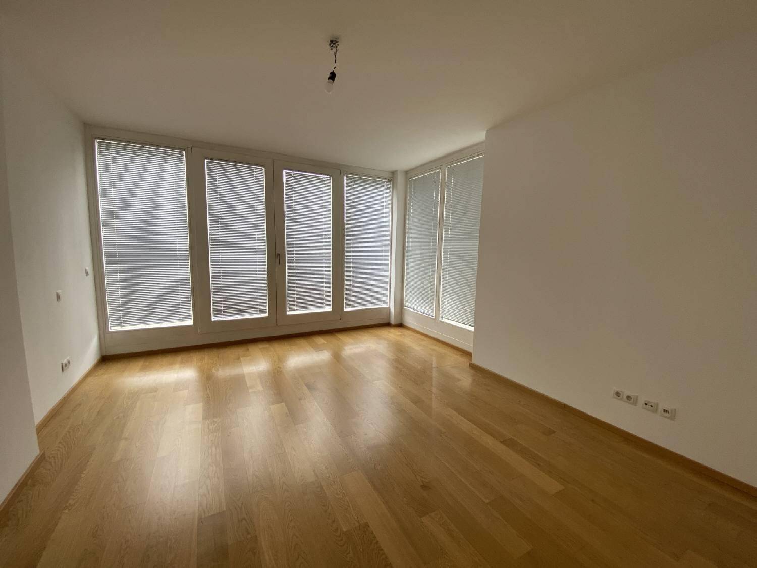 Leerer Raum mit Parkettboden und großen Fenstern, die mit Jalousien ausgestattet sind.