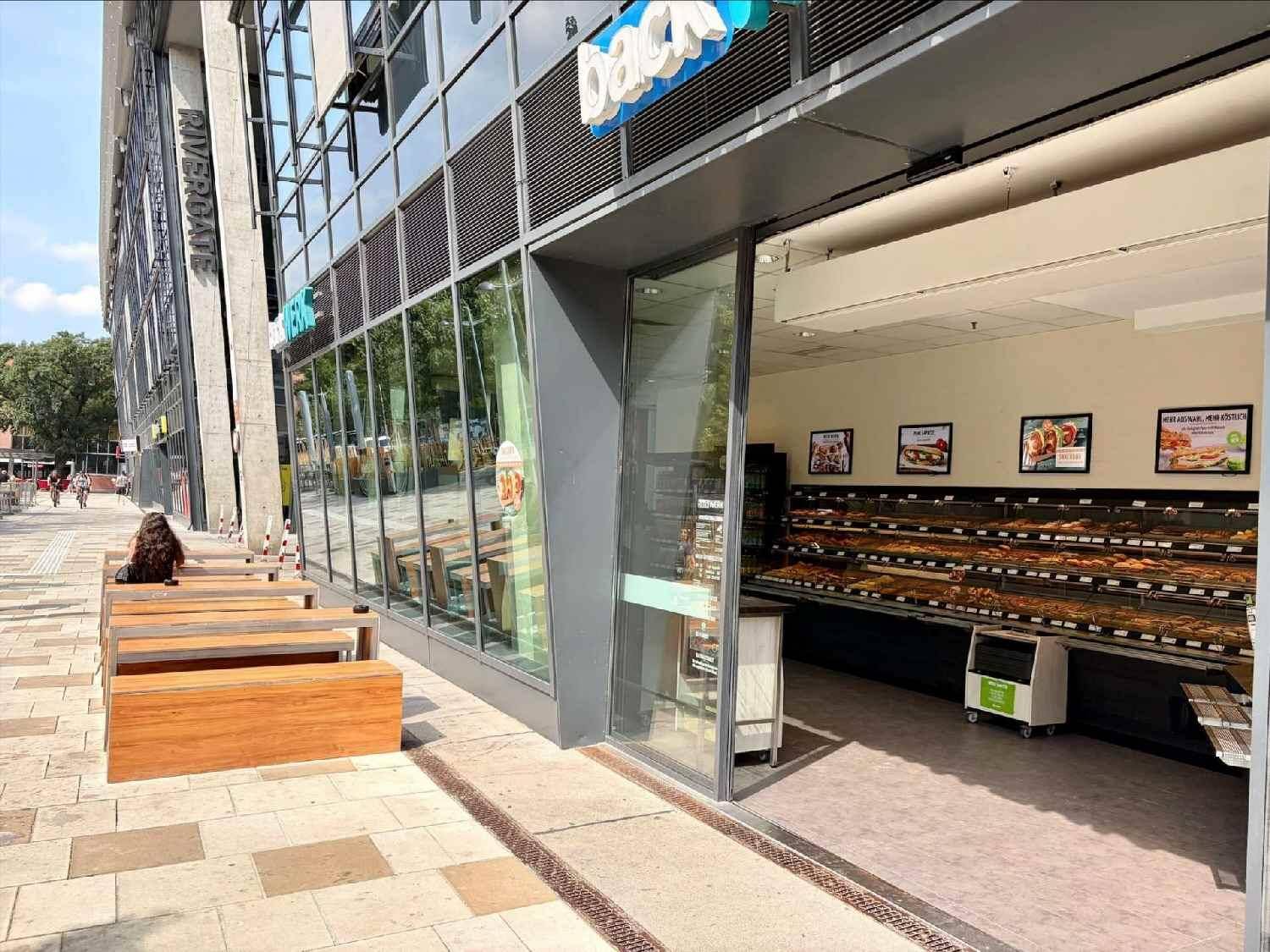 Eingangsbereich eines Geschäfts mit Auslagen und Sitzgelegenheiten im Freien an einem sonnigen Tag.