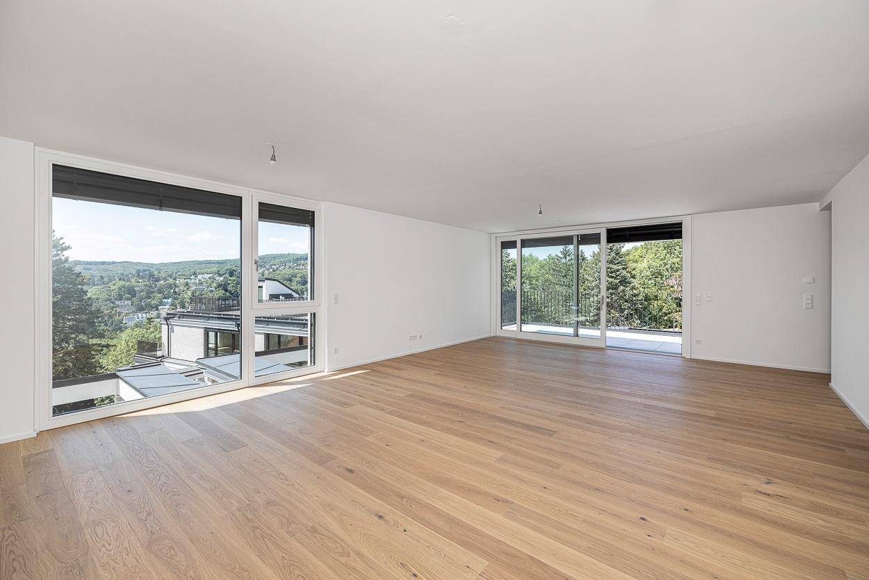 Heller, leerer Wohnraum mit großen Fenstern, Holzboden und weitem Blick ins Grüne.