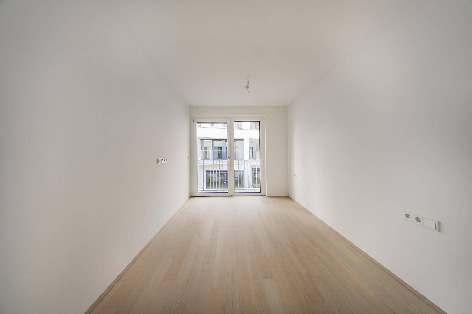 Leeres Zimmer mit hellem Holzboden und einem Fenster mit französischem Balkon.