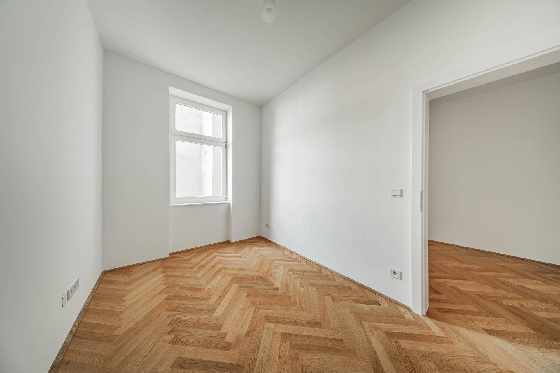 Lichtdurchfluteter Raum mit elegantem Fischgrätparkett und einem Fenster, das viel Tageslicht hereinlässt.