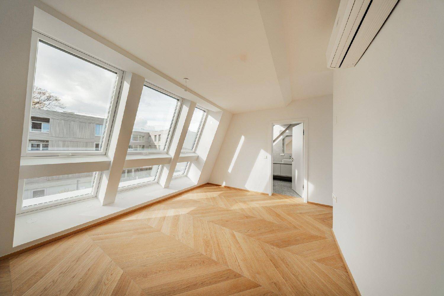 Helles Dachgeschosszimmer mit großen Fenstern, Fischgrätparkett und Blick in ein angrenzendes Badezimmer.
