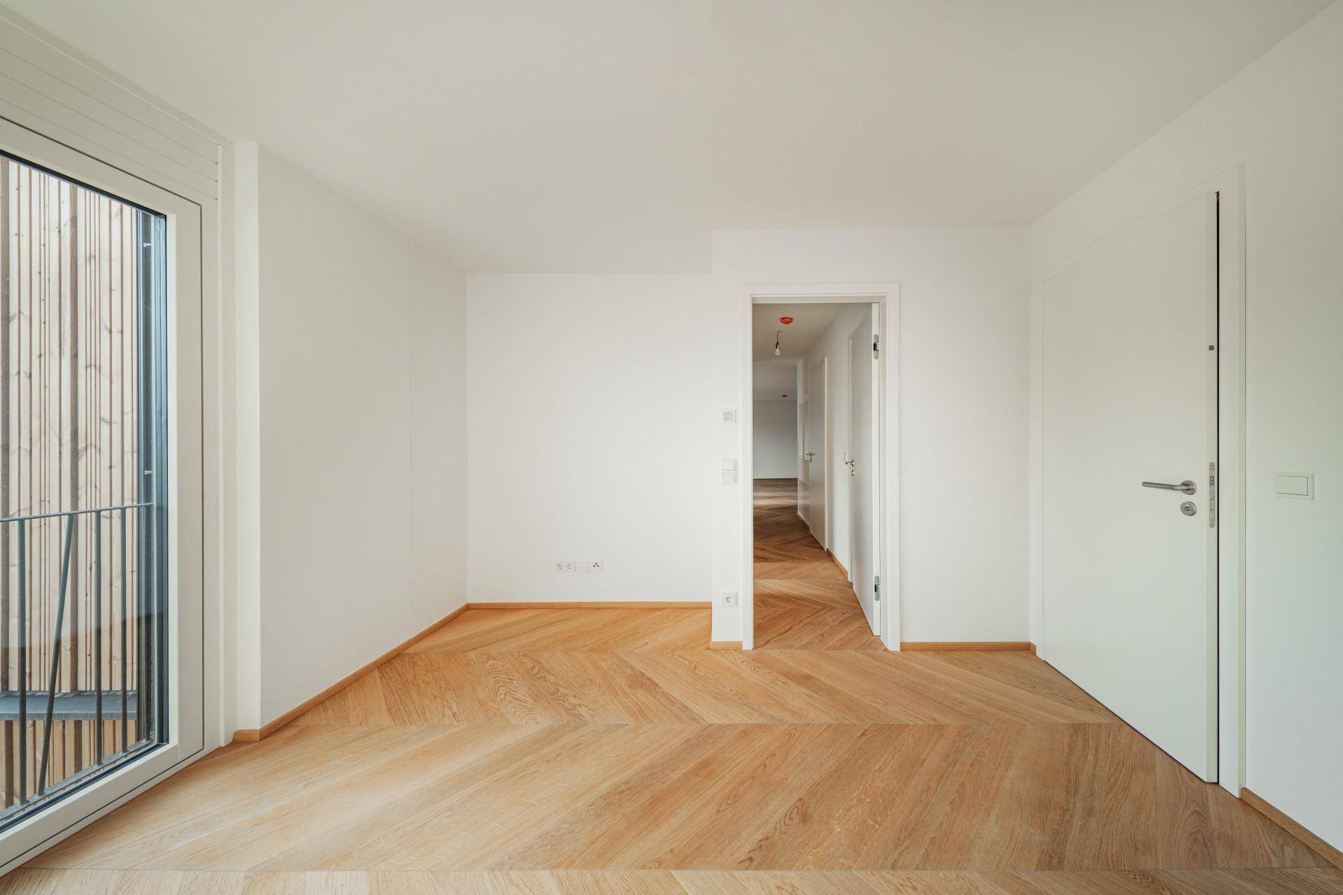 Helles Zimmer mit Fischgrätparkett und Zugang zu einem Balkon mit Holzverkleidung.