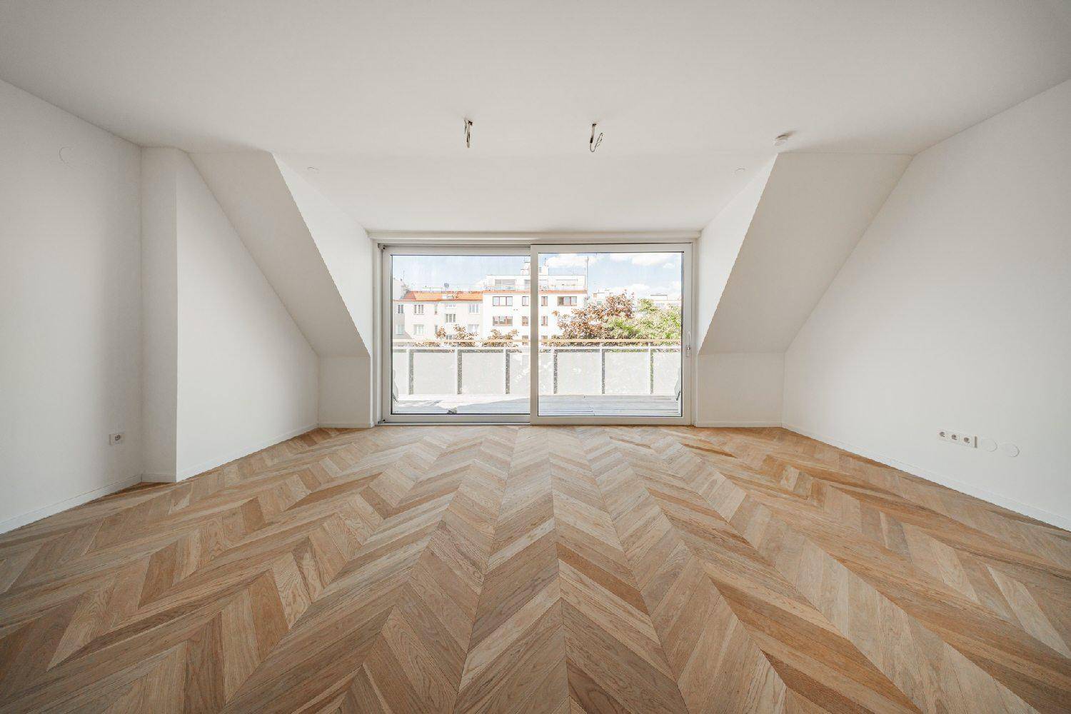 Heller Dachgeschossraum mit Fischgrätparkett, schrägen Wänden und Zugang zu einem Balkon.