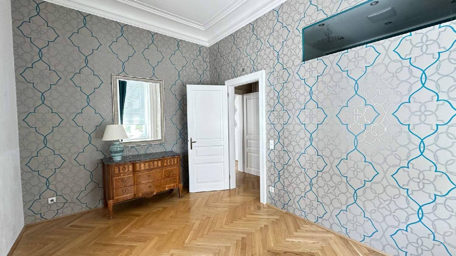 Zimmer mit gemusterten Tapeten, Fischgrätparkett und einem antiken Sideboard mit Spiegel.