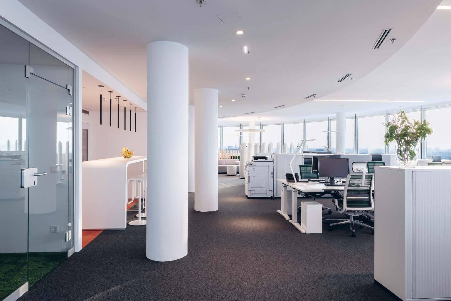 Offener Bürobereich mit Arbeitsplätzen, weißen Säulen und einem Empfangstresen, ausgestattet mit Teppichboden.