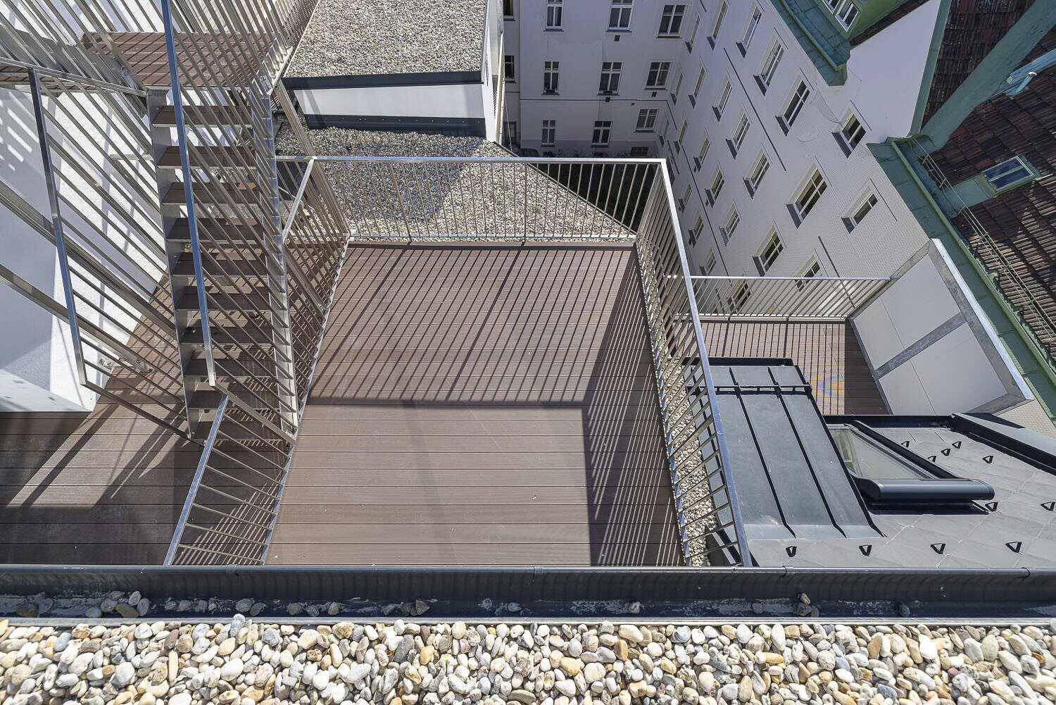 Dachterrasse mit Holzboden und Metallgeländer, umgeben von anderen Gebäuden und Kiesflächen.
