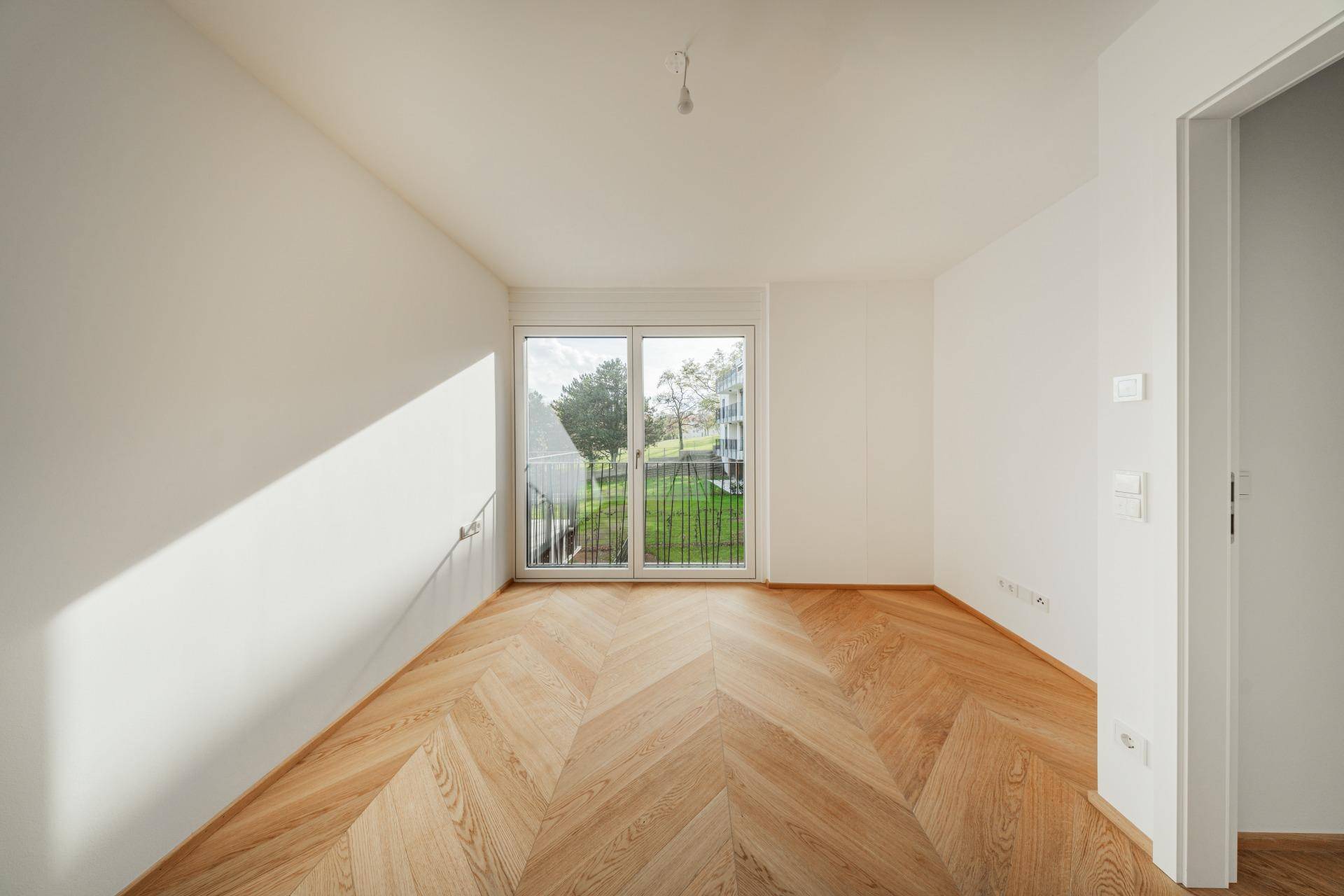 Helles Schlafzimmer mit Fischgrätparkett und Blick ins Grüne durch die Balkontür.