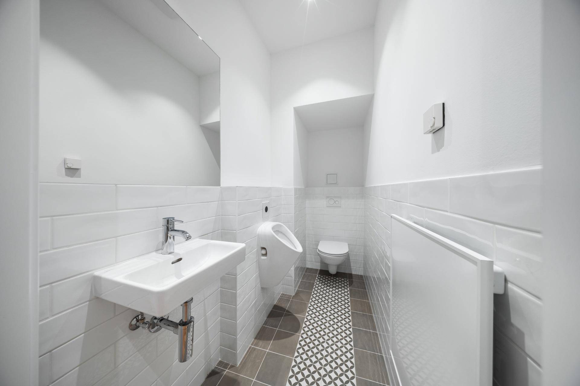 Modernes Gäste-WC mit weißen Fliesen, Waschbecken, Urinal und WC, sowie gemusterten Bodenfliesen.