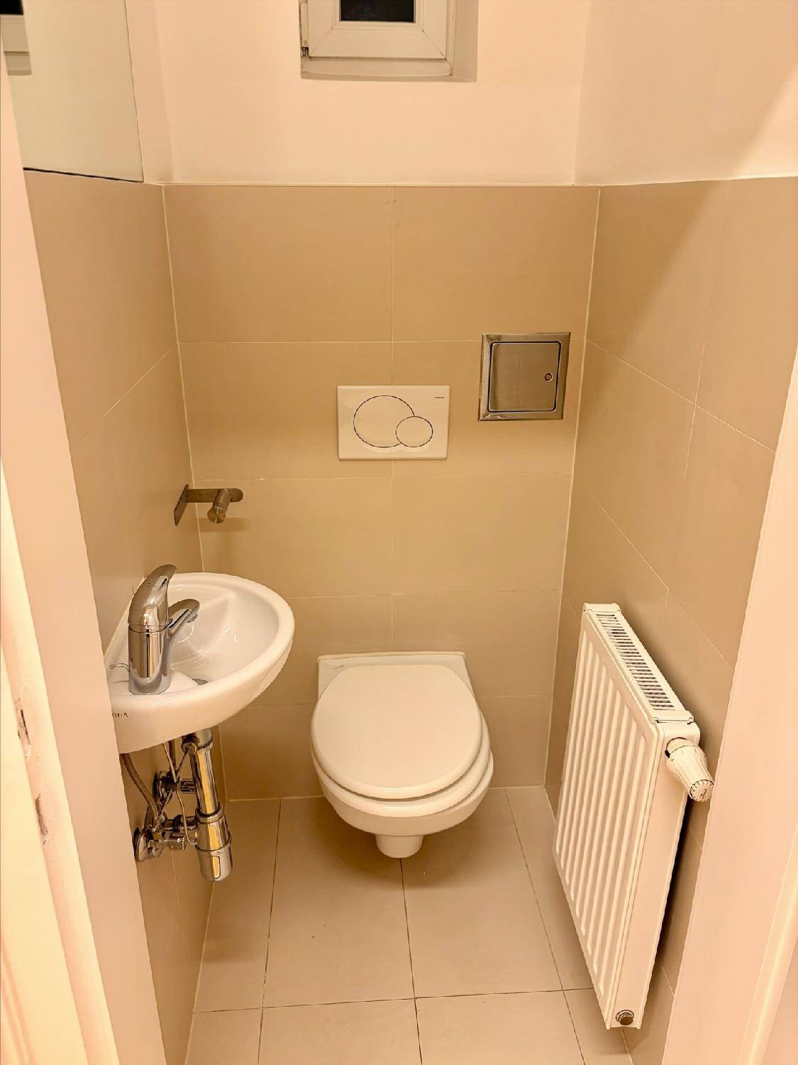 Kleiner Toilettenraum mit wandhängendem WC, Waschbecken, Heizkörper und beigefarbenen Fliesen.