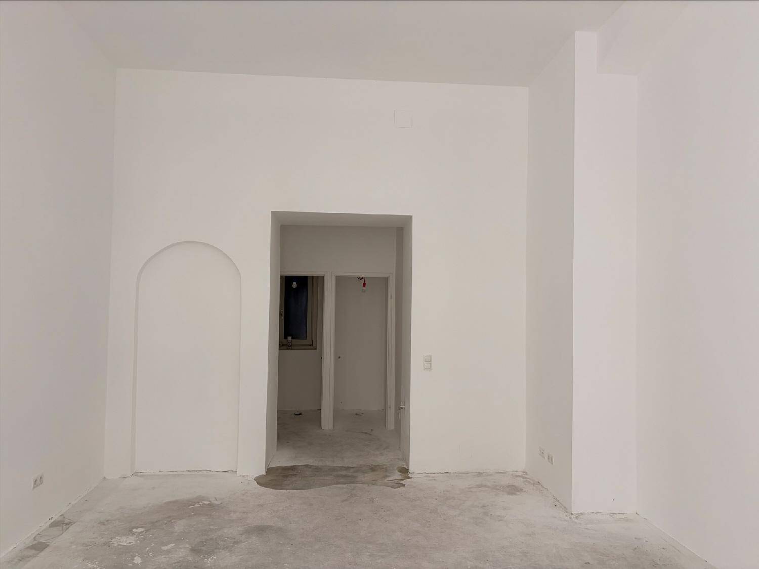 Leerer Raum mit weißen Wänden und Betonboden, mit einem Bogendurchgang und Tür zu weiteren Räumen.