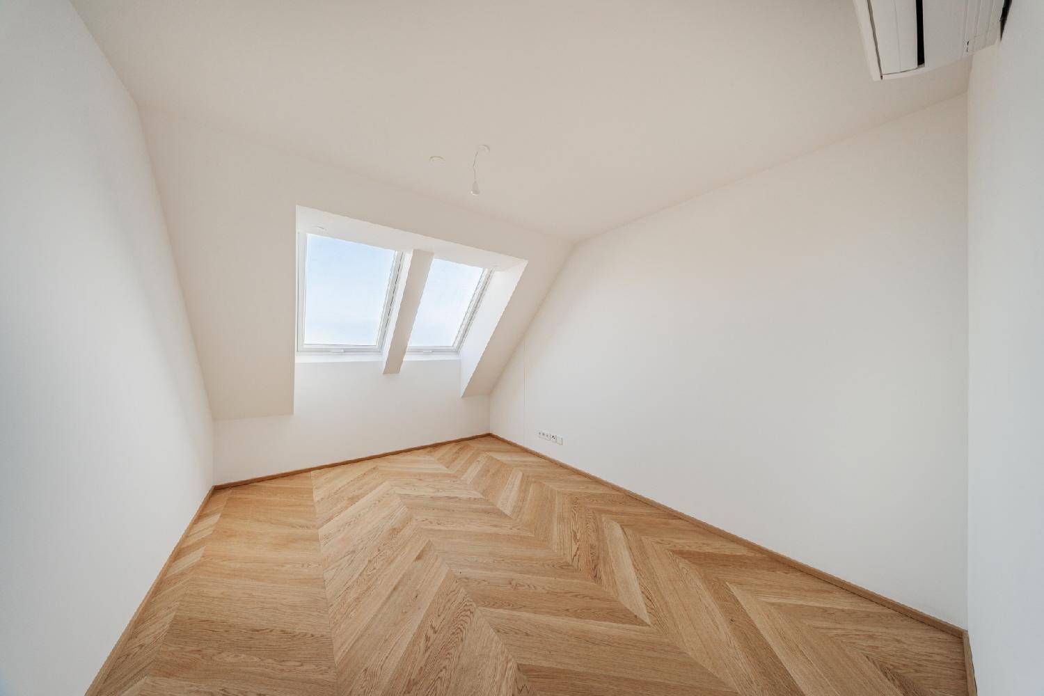 Helles Zimmer im Dachgeschoss mit schräger Decke, Parkettboden und zwei Dachfenstern.