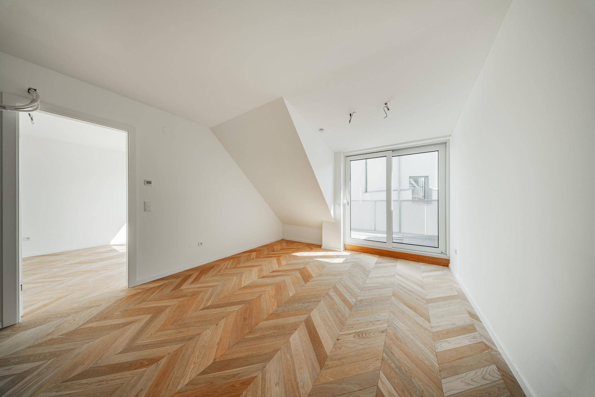 Heller Raum mit Fischgrätparkett, Dachschräge und Zugang zu einem kleinen Balkon.