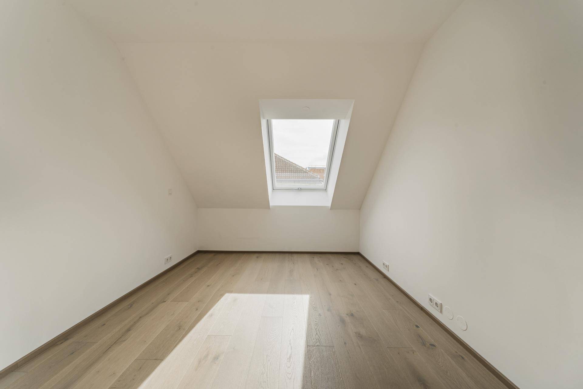 Geräumiges Zimmer mit Holzboden und Dachschräge, durch ein großes Dachfenster belichtet.