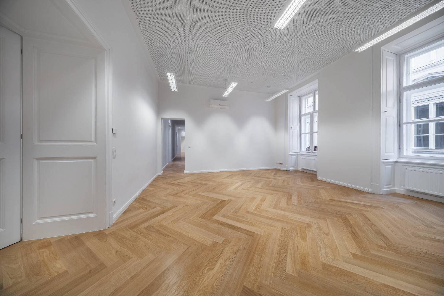 Geräumiger, heller Raum mit Fischgrätparkett, hohen Fenstern und einer Klimaanlage an der Wand.