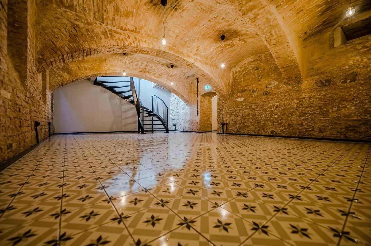 Historischer Gewölbekeller mit freiliegenden Ziegelwänden, gemusterten Fliesen und einer Wendeltreppe.