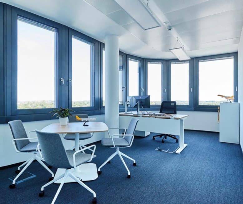Helles Eckbüro mit großen Fenstern, blauer Teppichboden und modernen Büromöbeln für eine angenehme Arbeitsatmosphäre.