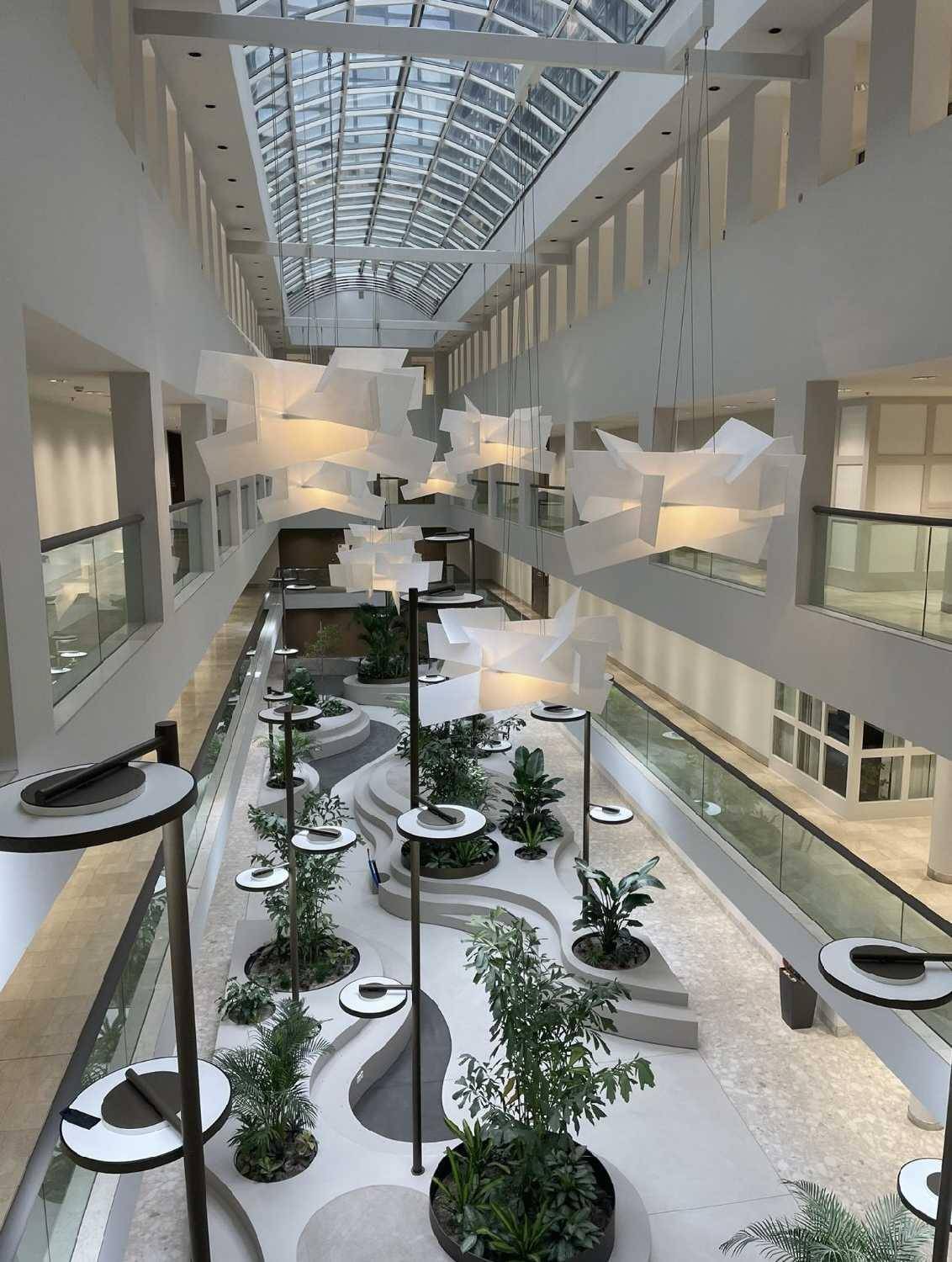 Geräumiges Atrium mit beeindruckendem Glasdach, modernen Leuchten und üppiger Innenraumbegrünung.