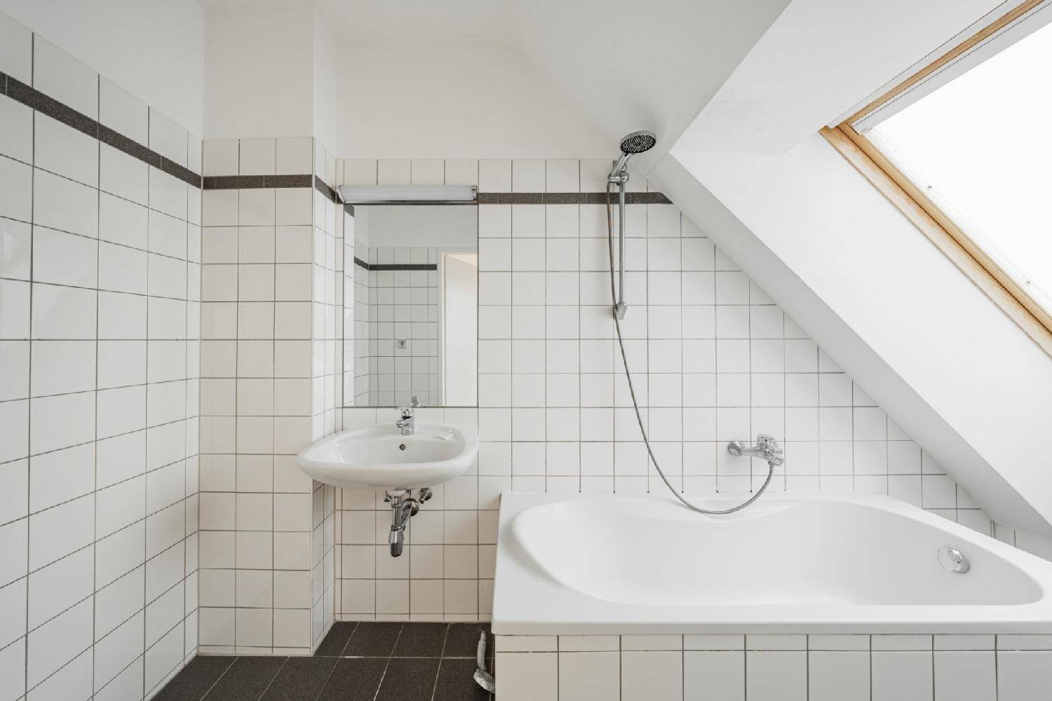 Badezimmer mit weißen Fliesen, Badewanne mit Duschfunktion und einem Dachfenster unter der Schräge.