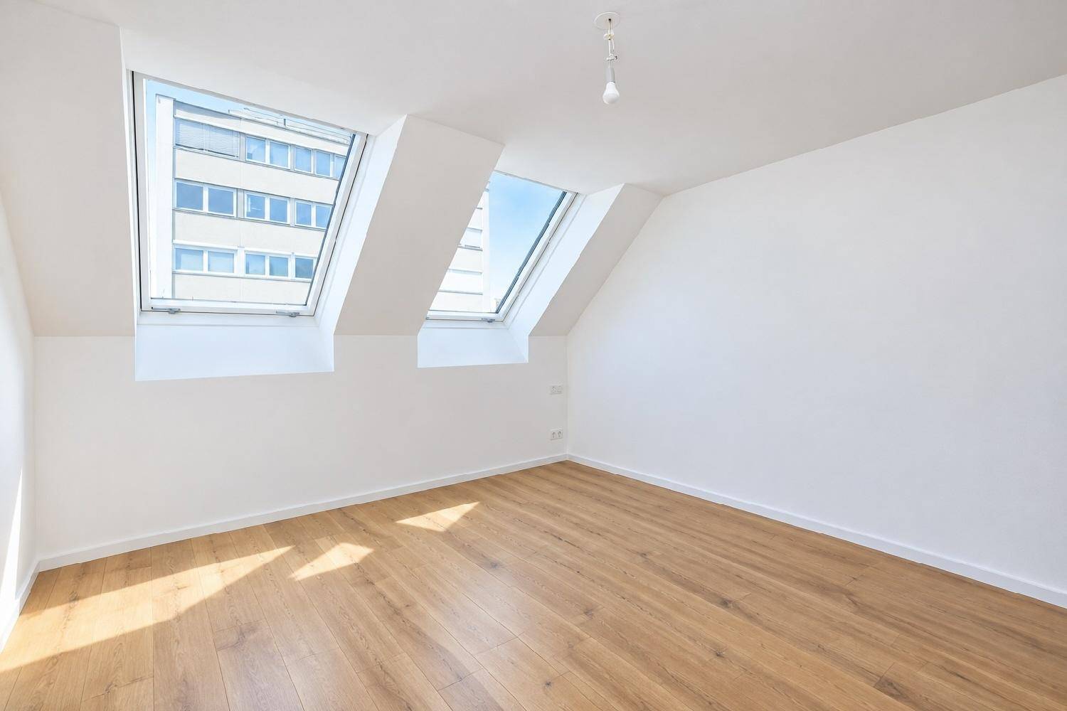 Lichtdurchflutetes Zimmer im Dachgeschoss mit Holzboden und Blick auf die Stadt durch Dachfenster.