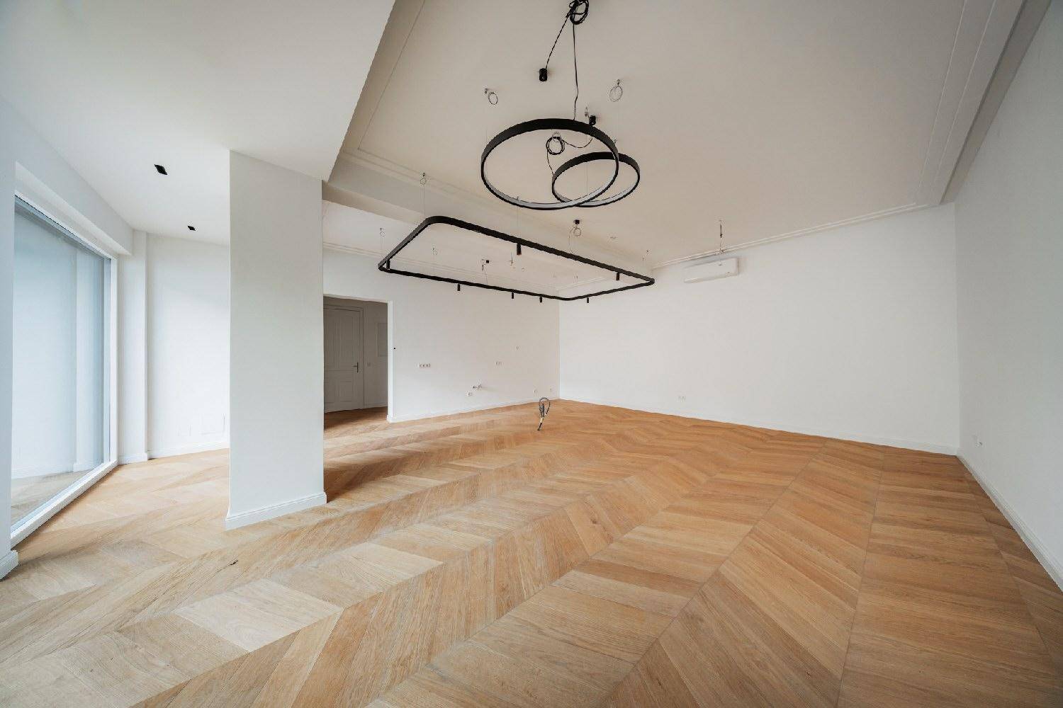 Großes, helles Zimmer mit Fischgrätparkett, modernen Leuchten und einer leichten Bodenerhöhung.