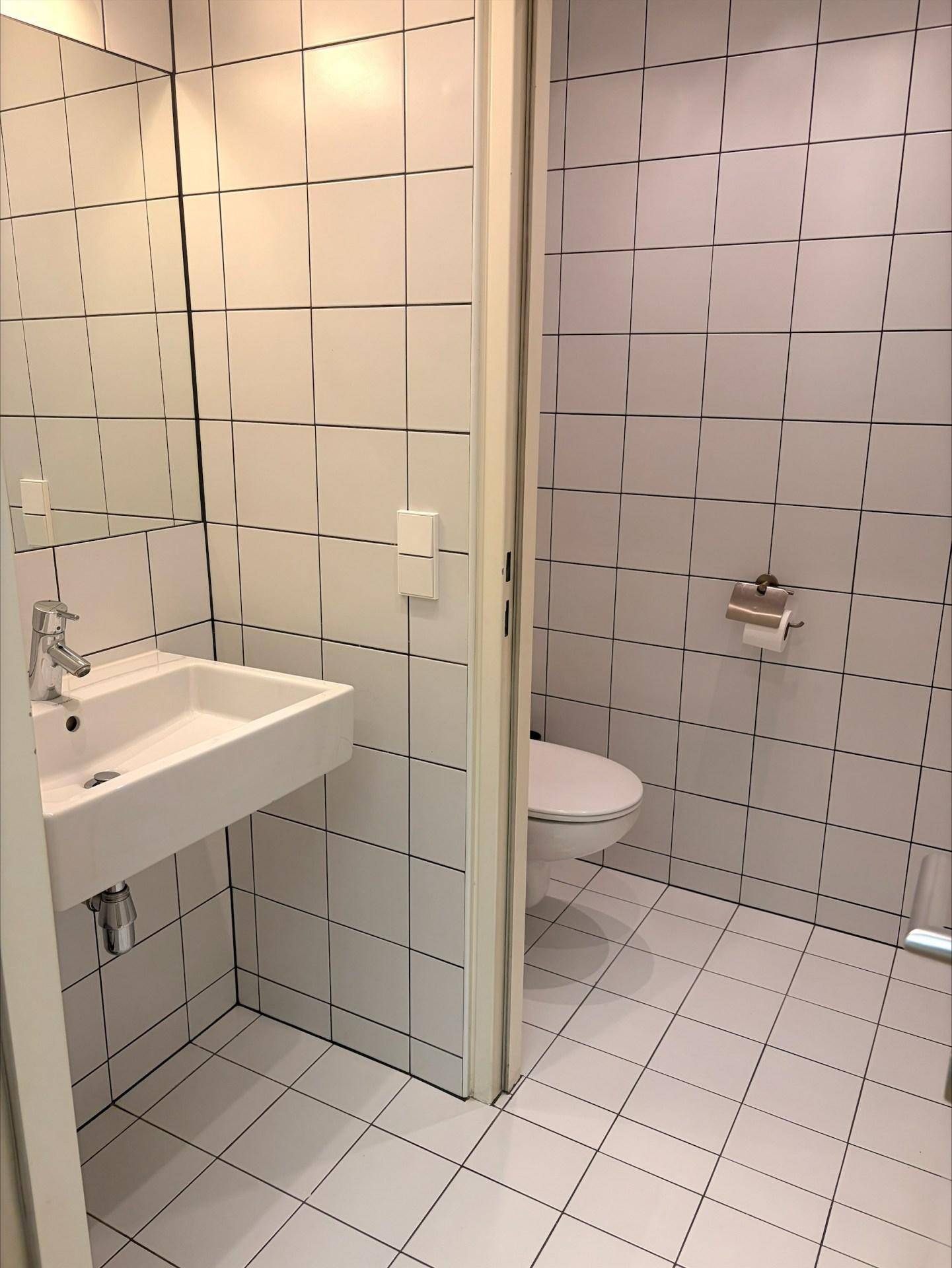 Modern gefliestes Badezimmer mit weißen Wand- und Bodenfliesen, einem Waschbecken und einer Toilette.