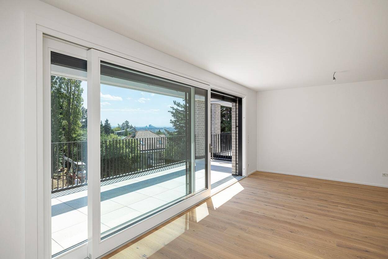Heller Raum mit Holzboden und großem Schiebefenster zum Balkon, mit Blick ins Grüne und auf die Stadt.