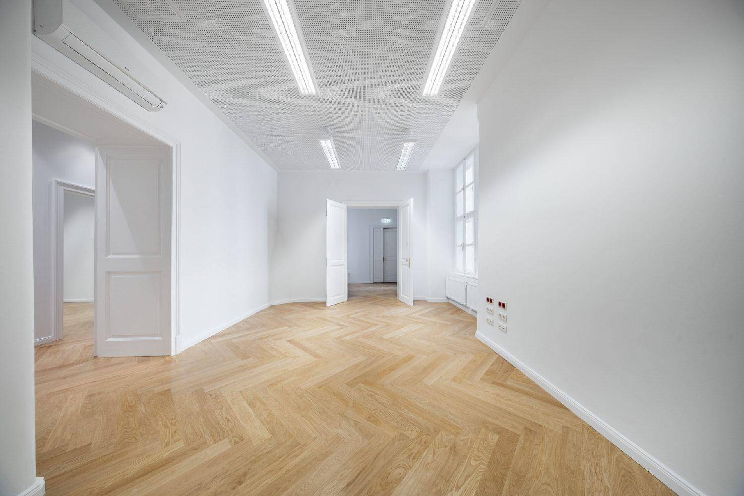Heller, leerer Raum mit Fischgrätparkett, weißen Wänden, Klimaanlage und großem Fenster.