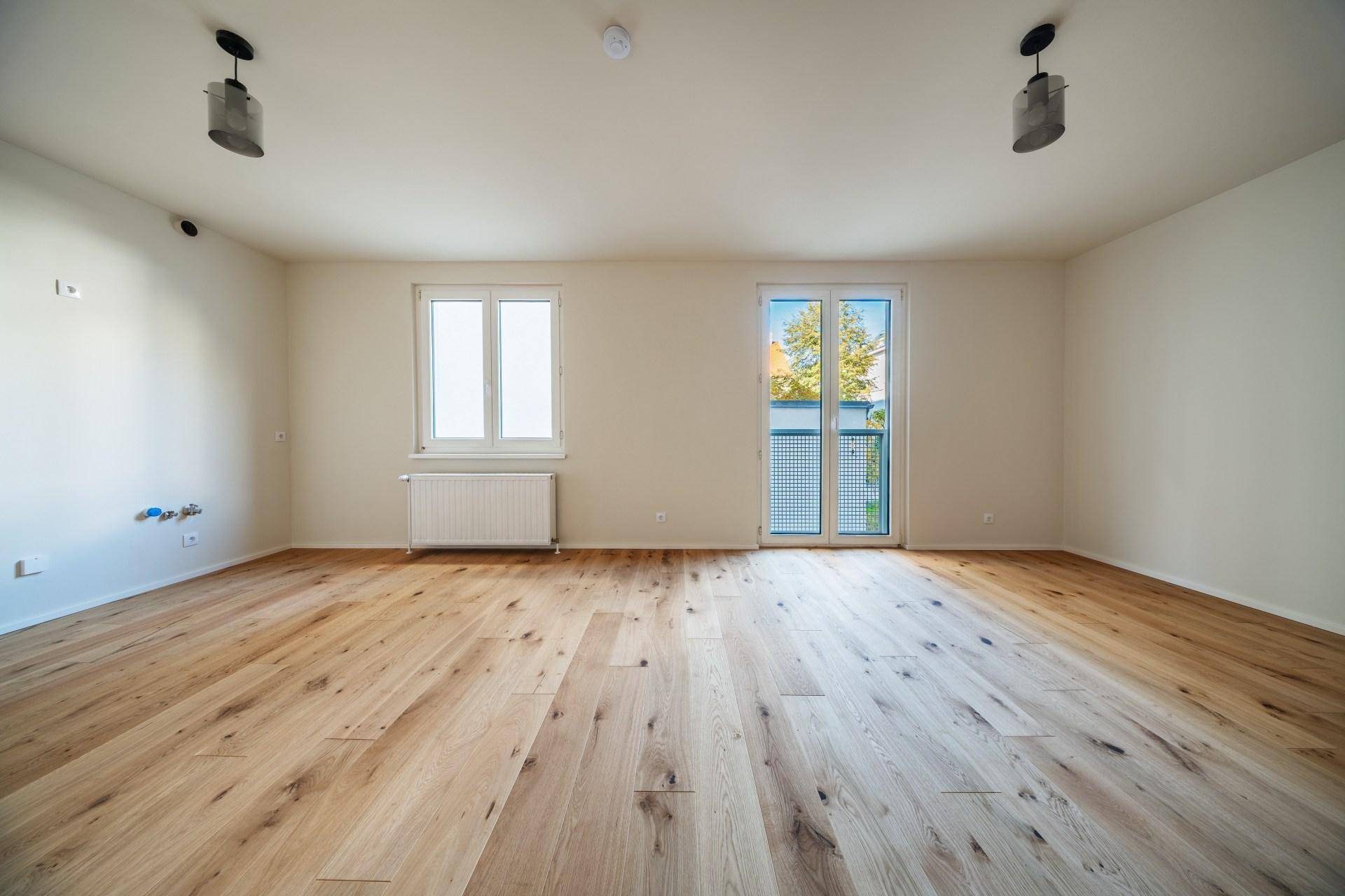 Geräumiges, helles Zimmer mit Holzboden, zwei Fenstern und einem Heizkörper unter einem Fenster.