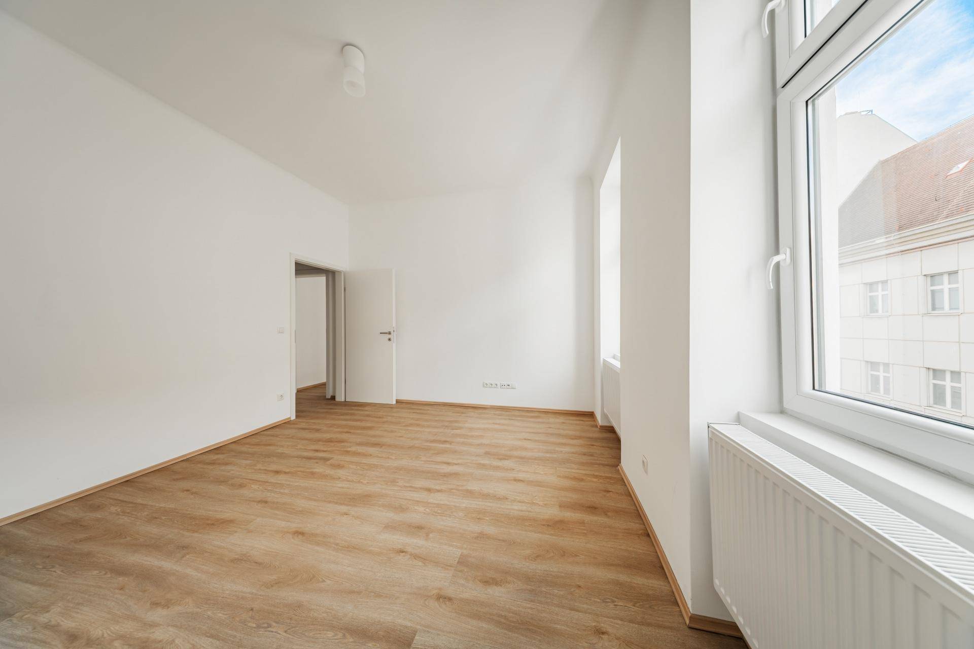 Geräumiges Wohnzimmer mit Laminatboden und großen Fenstern, die viel Tageslicht hereinlassen.