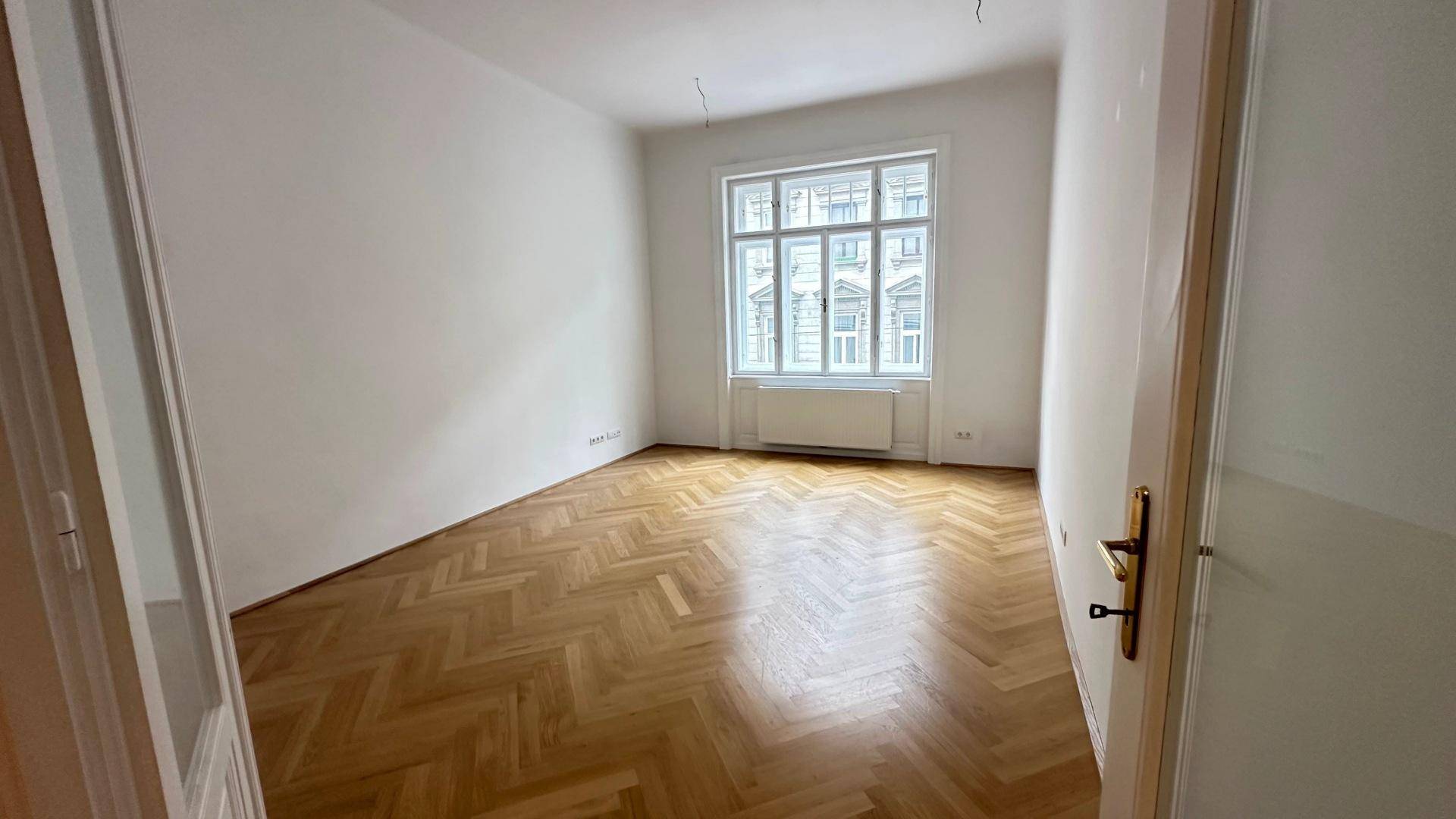 Geräumiges Zimmer mit klassischem Fischgrätparkett und einem großen Fenster, das viel Licht hereinlässt.