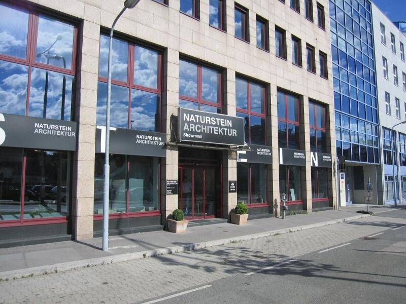 Eingangsbereich eines modernen Geschäftsgebäudes mit großen Glasfronten, die den Himmel widerspiegeln, und Beschilderung für ein Architekturbüro.