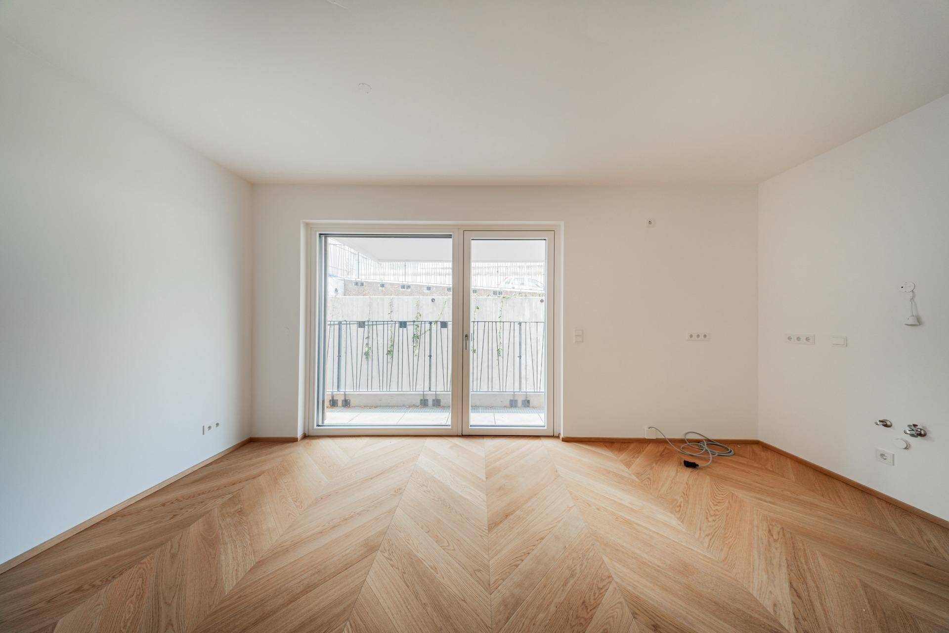 Geräumiger, heller Raum mit Fischgrätparkett und breiter Glasfront zum Balkon.