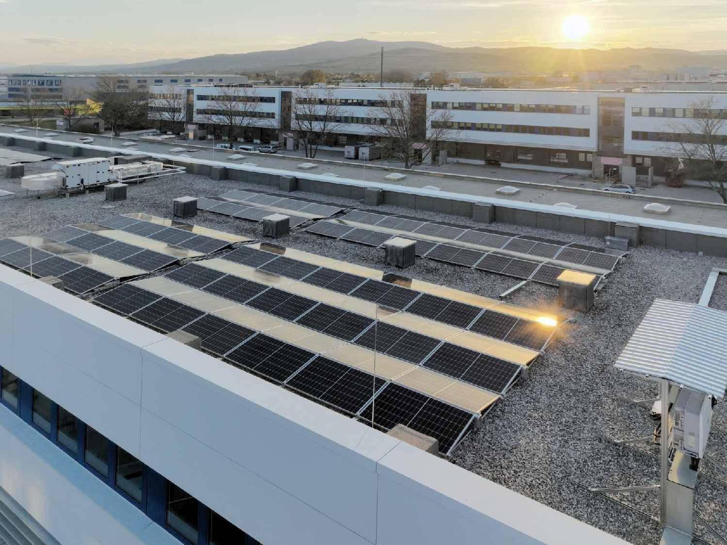 Dachansicht eines Gebäudes mit zahlreichen Photovoltaik-Modulen bei Sonnenuntergang und Bergblick.