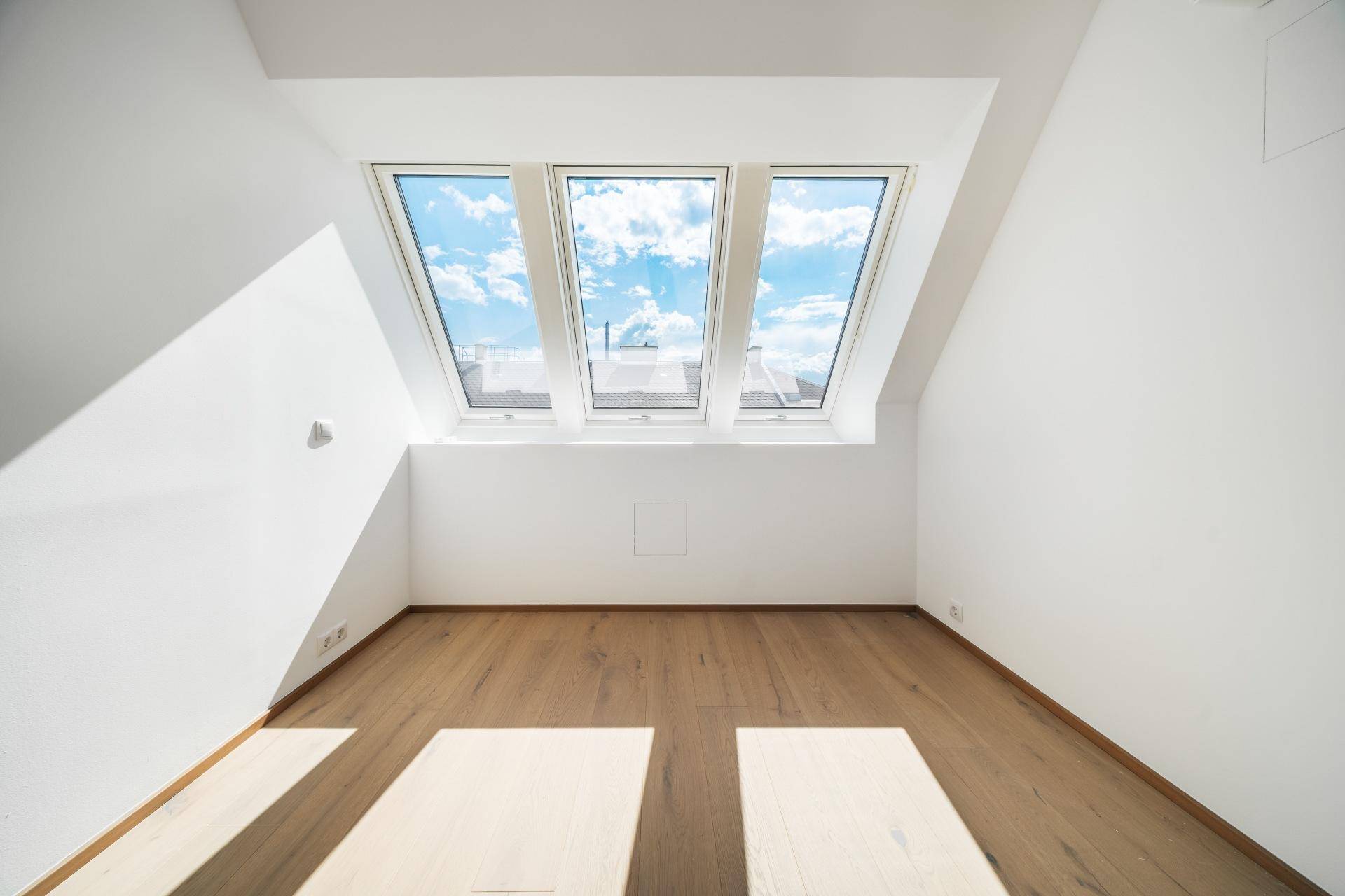 Helles Schlafzimmer mit Holzboden und drei großen Dachfenstern, die viel Tageslicht hereinlassen.