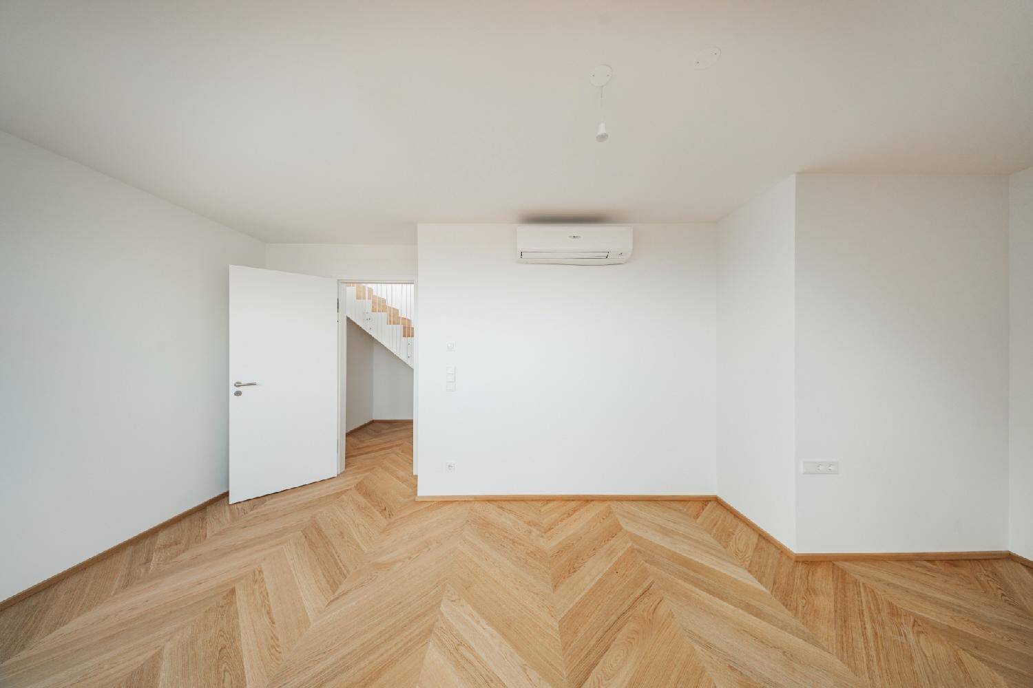 Leeres Zimmer mit weiß gestrichenen Wänden, Fischgrätparkett und Zugang zum Treppenhaus.