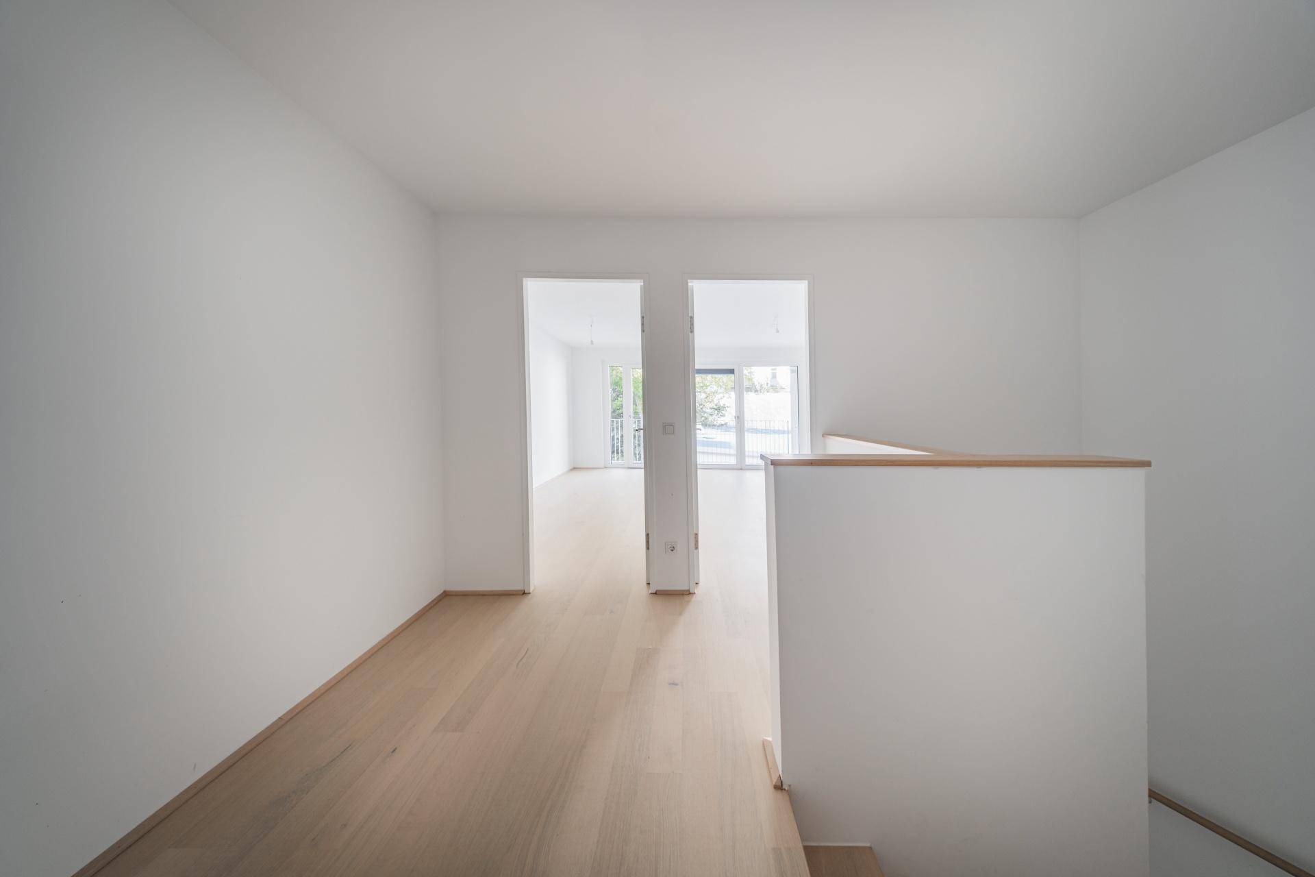 Offener Wohnbereich mit hellem Holzboden und Blick auf einen weiteren Raum sowie eine Treppe.