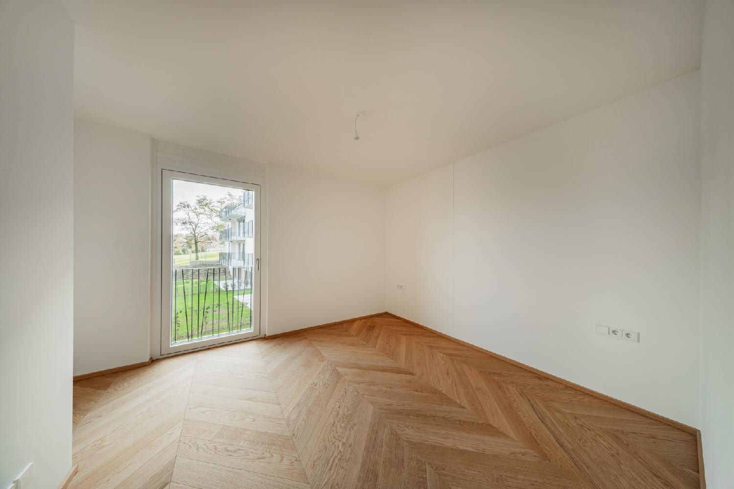 Freundliches Zimmer mit Fischgrätparkett und großem Fenster, das viel Tageslicht hereinlässt.