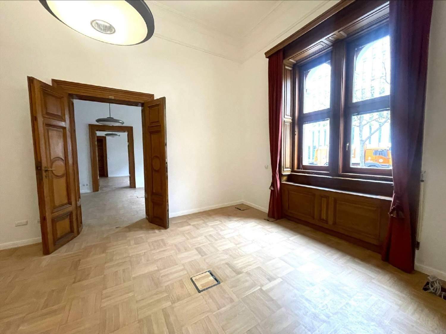 Großes Holzfenster mit roten Vorhängen in einem hellen Raum mit klassischem Fischgrätparkett und hohen Decken.