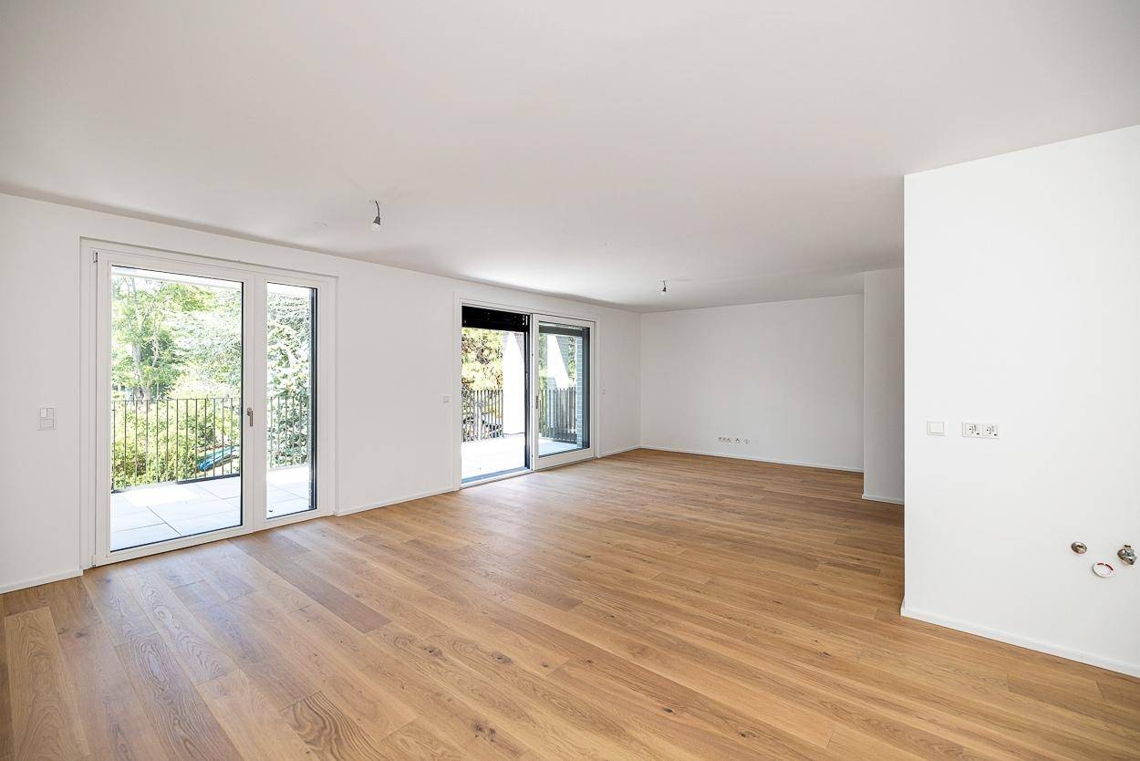 Geräumiger, leerer Wohnbereich mit hellem Holzboden und Zugang zu zwei Balkonen oder Terrassen.