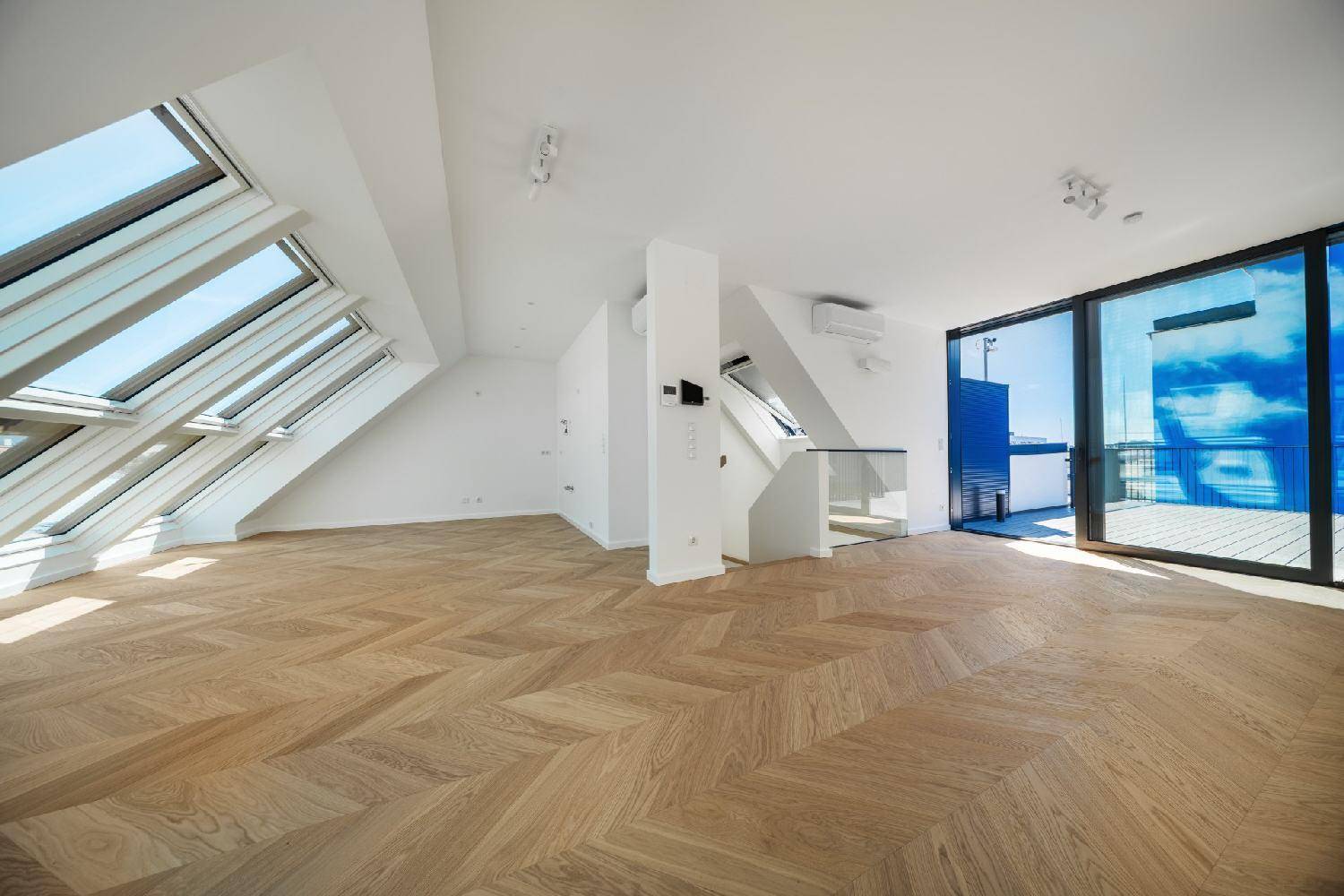 Heller Wohnraum mit elegantem Fischgrätparkett, Klimaanlage und modernem Glasgeländer, das den Treppenbereich abgrenzt.